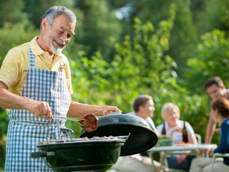 Older man cooking at BBQ with family in the background