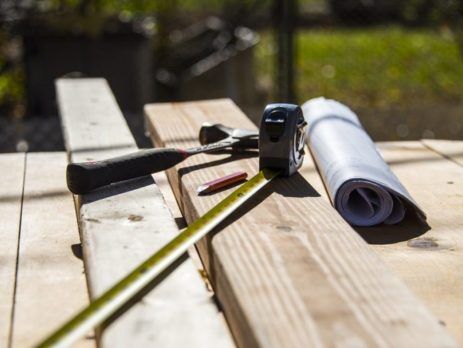 House plans, hammer and measuring tape sitting on wooden deck surface under construction